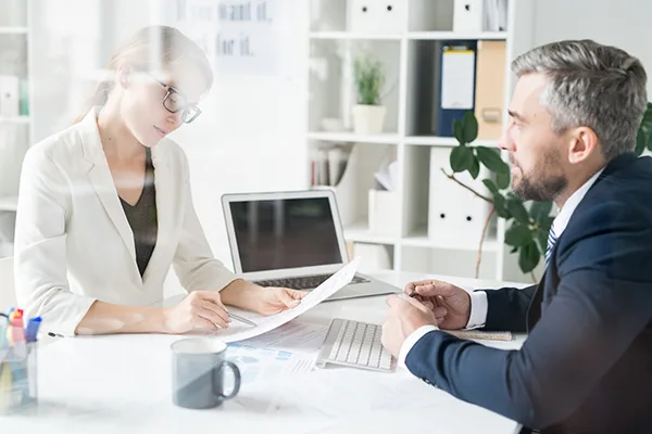 Serious concentrated female financial adviser in glasses sitting at desk and analyzing papers with businessman while giving advises about strategy and approach to him